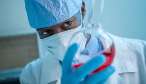 A medical scientist in protective garments and mask is holding up and inspecting a glass vase with red liquid content.