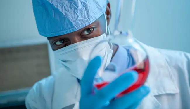 A medical scientist in protective garments and mask is holding up and inspecting a glass vase with red liquid content.